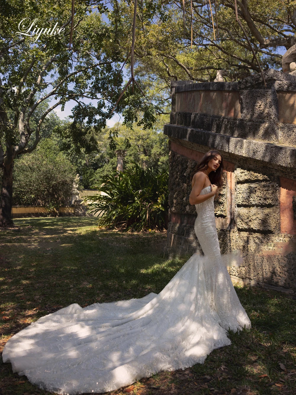 Stately ivory lace mermaid bridal dress on a model next to an outdoor stone staircase, displaying long embroidered sleeves with statement feather accents and a flared skirt.