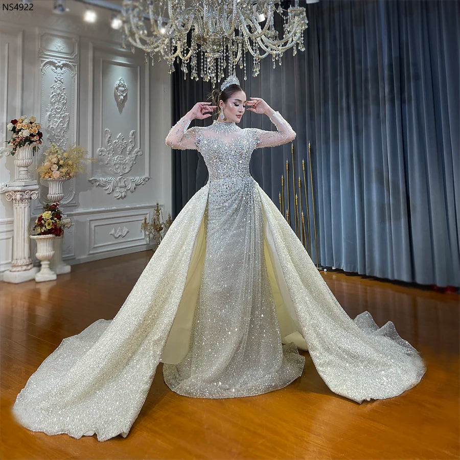 A bride wearing a luxury 2-in-1 wedding dress featuring a high neckline, long sheer sleeves, and a dramatic detachable overskirt, all heavily embellished with shimmering crystals and pearls.
