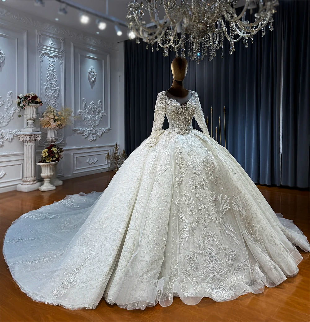Elegant front-facing view of a long-sleeve bridal ball gown on a mannequin, showcasing the intricate silver beadwork, shimmering fabric, and majestic cathedral train under a crystal chandelier.