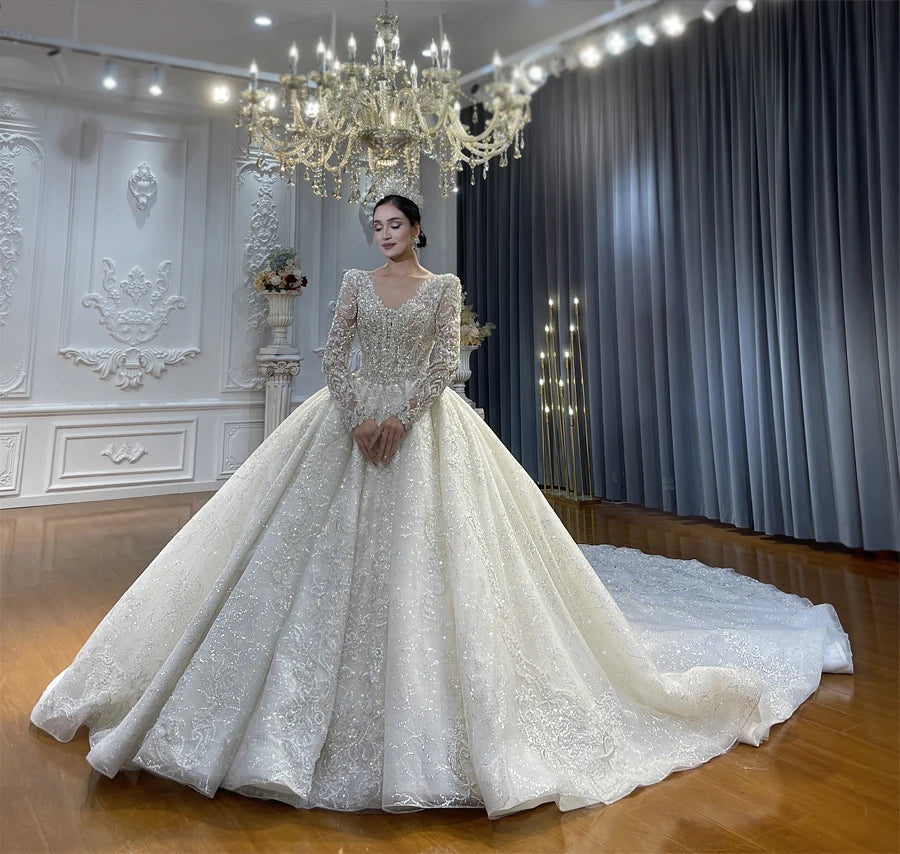 Elegant long-sleeved wedding dress under a chandelier, showcasing the removable beaded neckline and the shimmering, intricate lace detailing throughout the gown.