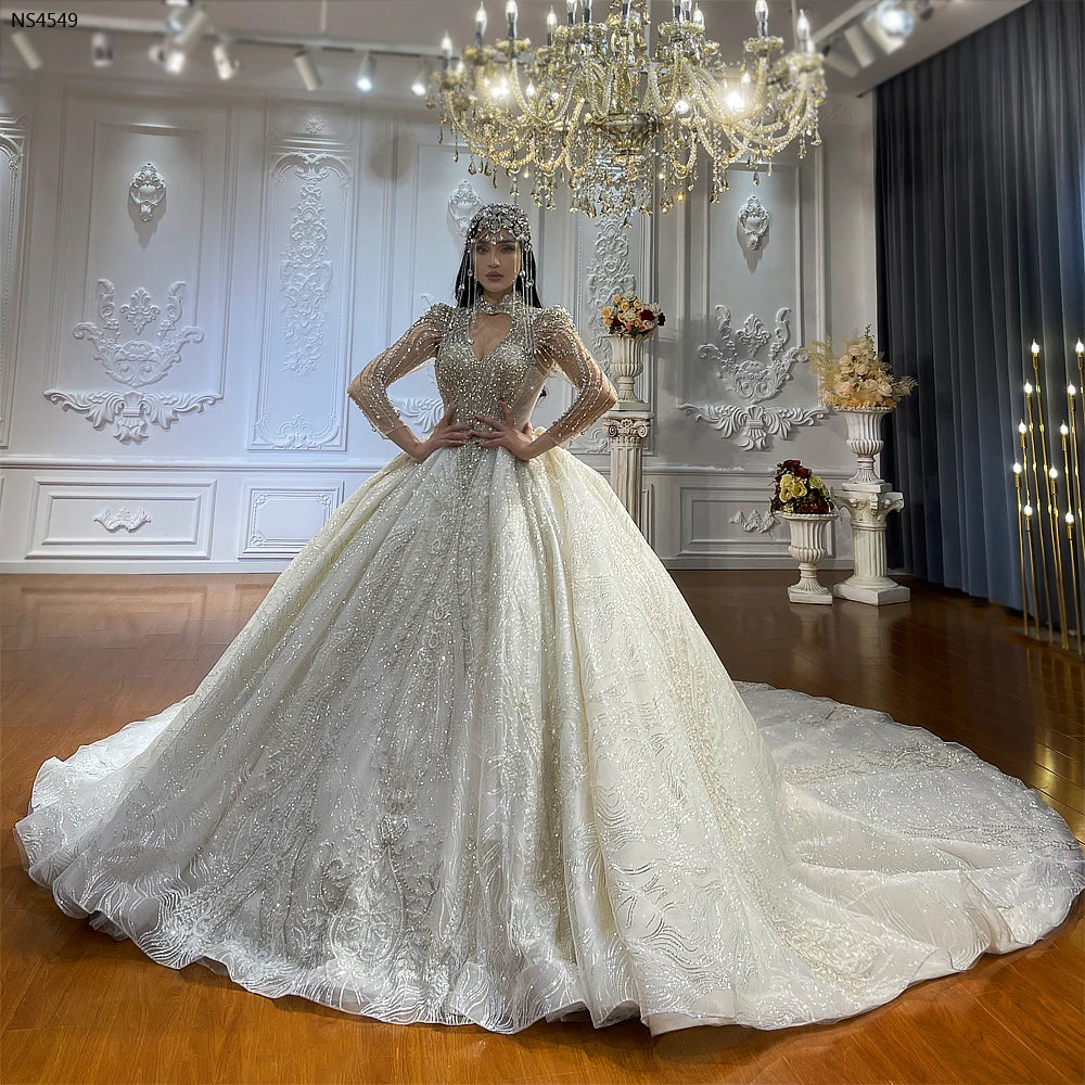 Full-length front view of a bride wearing a luxury Dubai style wedding dress, featuring an expansive hand-beaded ball gown skirt, long sheer sleeves, and a matching crystal headpiece beneath an ornate chandelier.
