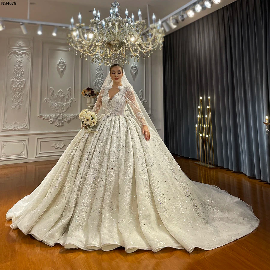 Full front view of a bride wearing a luxury Dubai wedding dress, an elegant long-sleeve ball gown bridal couture with a massive crystal-embellished train and sheer illusion neckline.