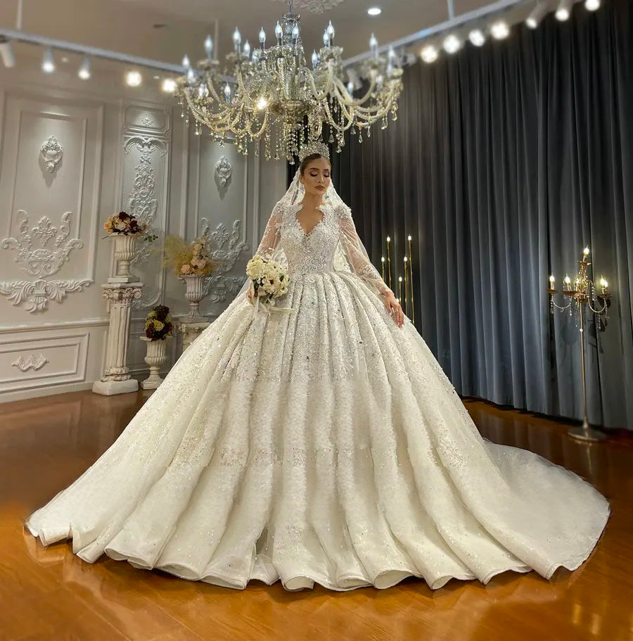A stunning bride models a luxury Dubai wedding dress, featuring a deep V-neck illusion bodice, long lace sleeves, and a heavy, shimmering ball gown skirt with a grand train.