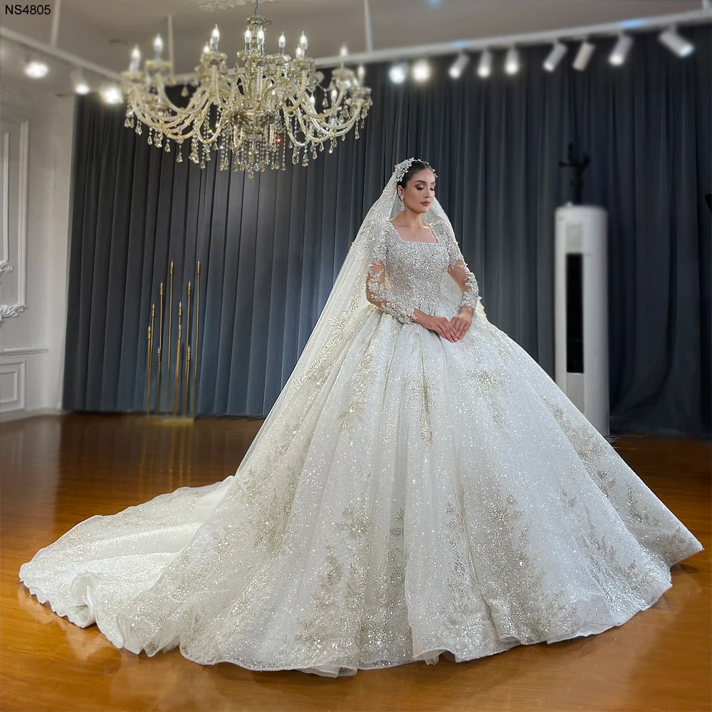A gorgeous bride showcasing a luxury handmade ball gown wedding dress featuring a modern square neckline, heavy silver beadwork, long sleeves, and a sparkling cathedral train.