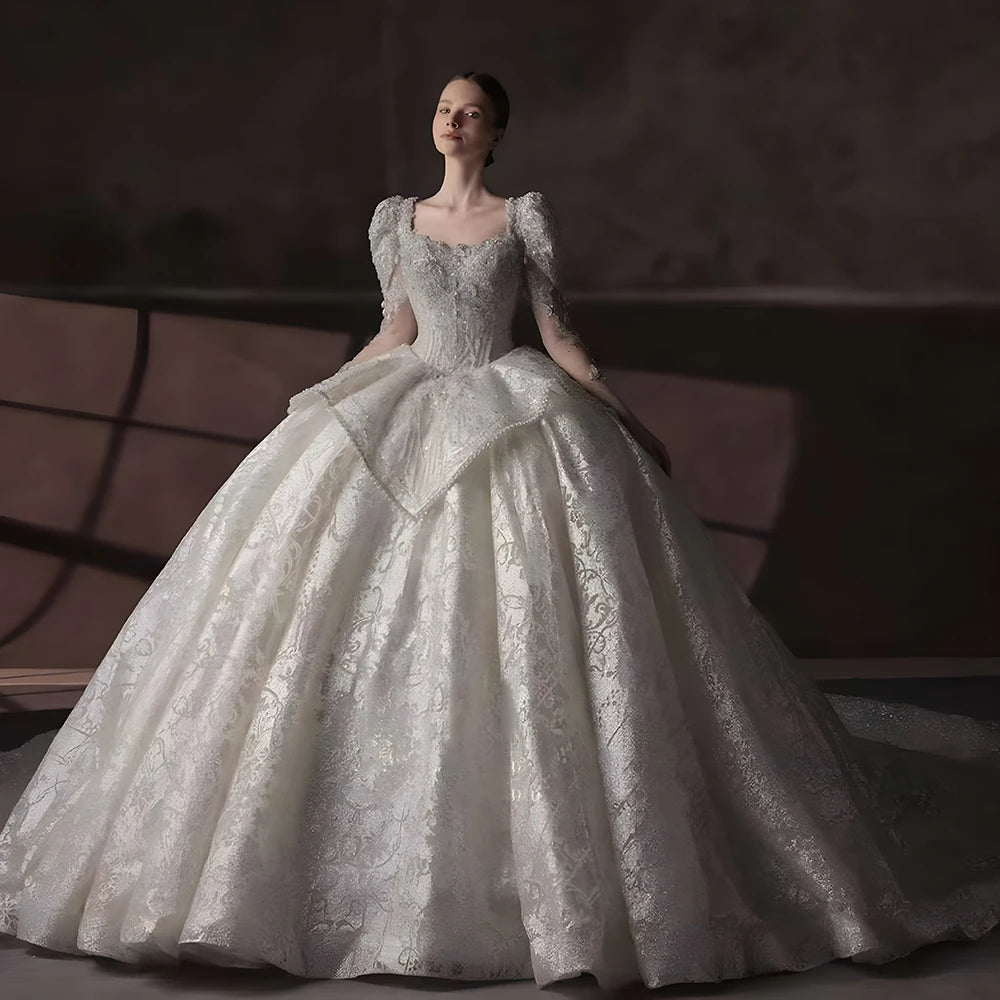 Full front view of a luxury international ball gown wedding dress featuring a square collar, long sleeves, and high-end shimmering fabric in a bridal showroom.