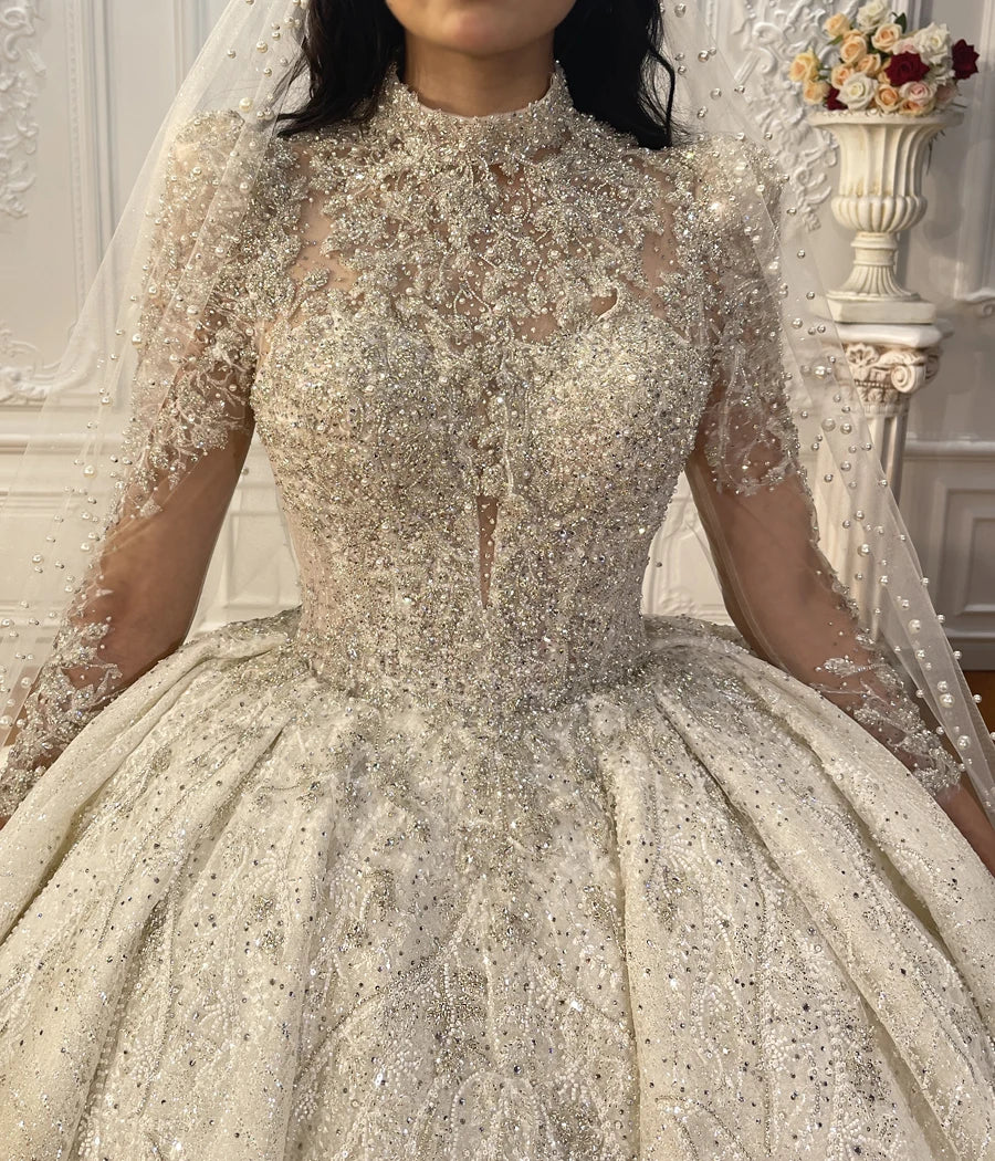 Close-up of the luxury wedding dress bodice, showcasing intricate heavy beadwork, pearl accents, a high illusion neckline, and elegant lace sleeves.