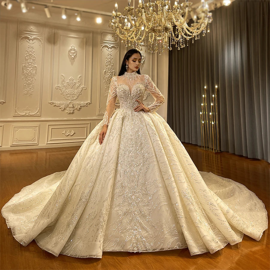 Front-side view of an elegant model wearing a luxury lace ball gown wedding dress with a high cover neck and sheer long sleeves, featuring intricate shimmering crystal embroidery and a voluminous skirt in a grand showroom.