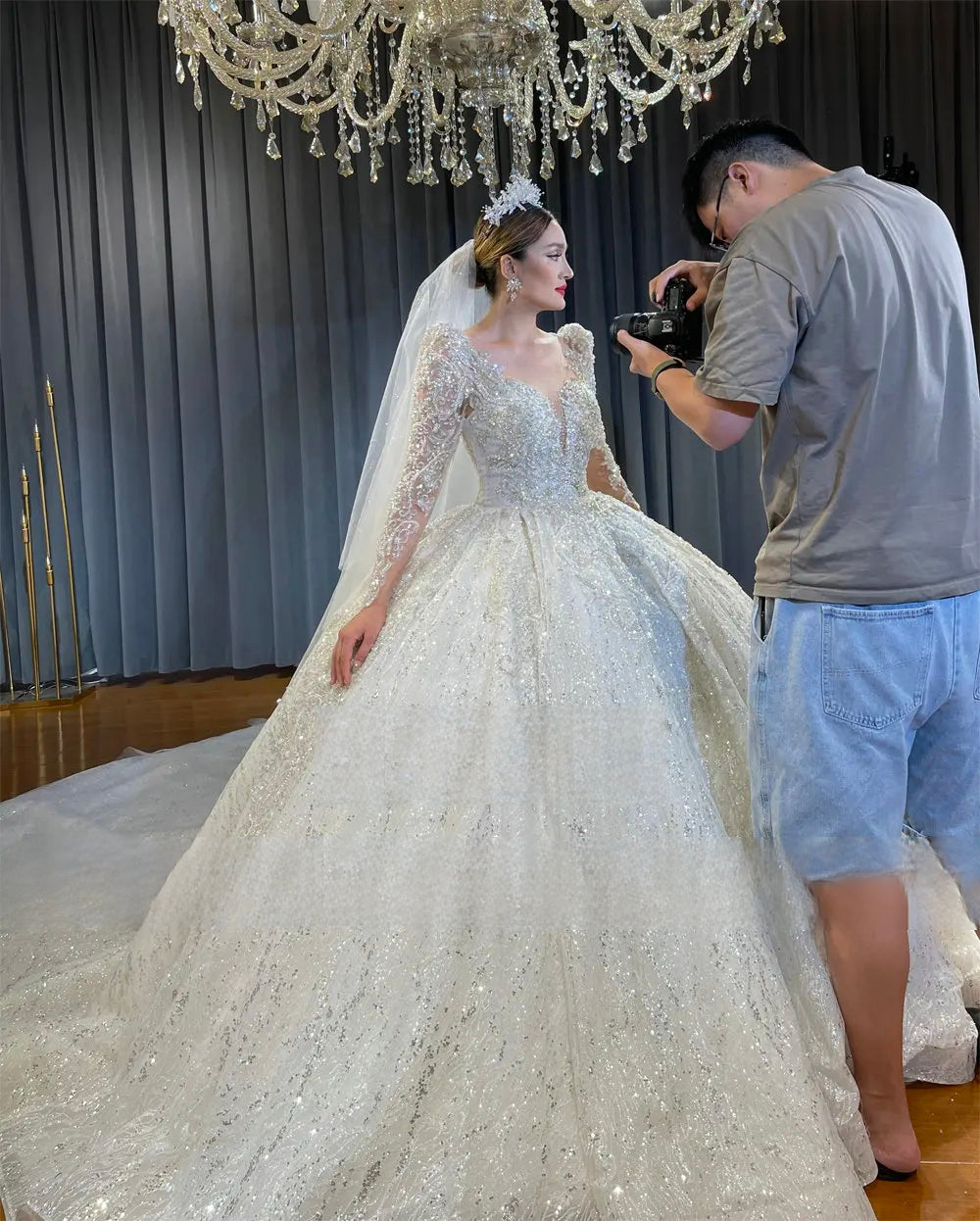 Side profile of a bride in a sparkling long sleeve crystal wedding dress with a low cut neckline and matching tiara, showcasing the beginning of a magnificent long train.