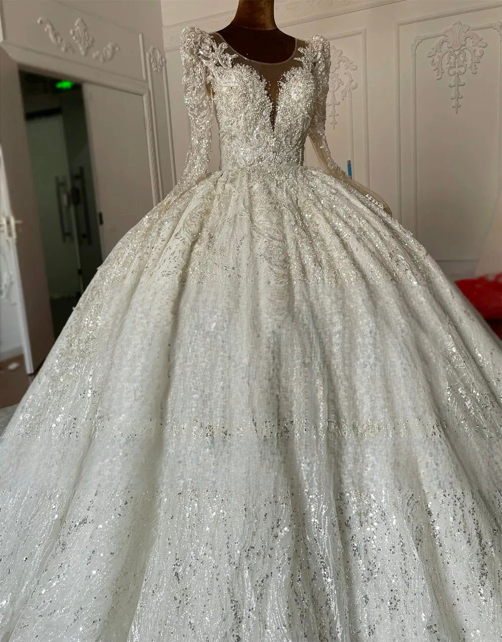 Close-up of a luxury bridal gown bodice on a mannequin, highlighting the detailed lace appliques, crystal embellishments, and long sleeves with a plunging illusion neckline.