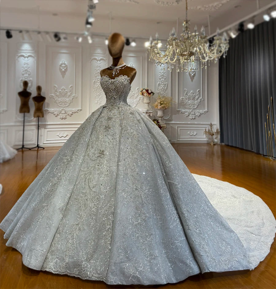 Stunning silver luxury ball gown wedding dress displayed on a mannequin, highlighting the heavy crystal beadwork, floral lace patterns, and a voluminous flared skirt with an extra-long cathedral train.