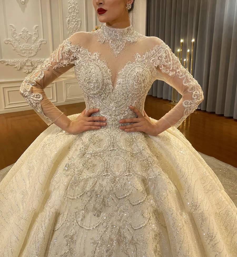 Frontal view of a luxury bridal gown on a mannequin, showcasing the intricate lace embroidery, pearl-beaded high neck, and the structured drape of the elegant long cape.