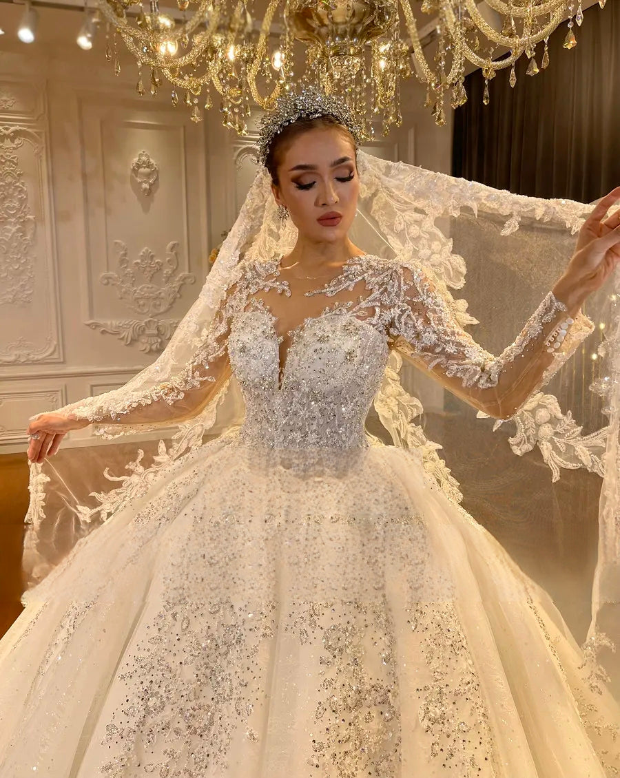 Close-up detail of an elegant ivory bridal gown bodice showcasing intricate lace appliques, hand-beaded sequins, and sheer long sleeves paired with a royal crystal tiara.