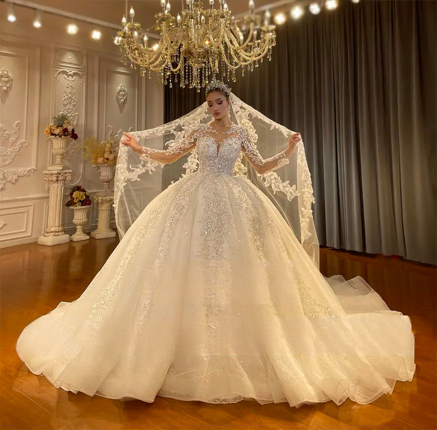Regal front perspective of a sparkly ball gown wedding dress with sheer lace sleeves and a voluminous skirt, displayed with a beautiful cathedral-length veil in a luxury bridal showroom.