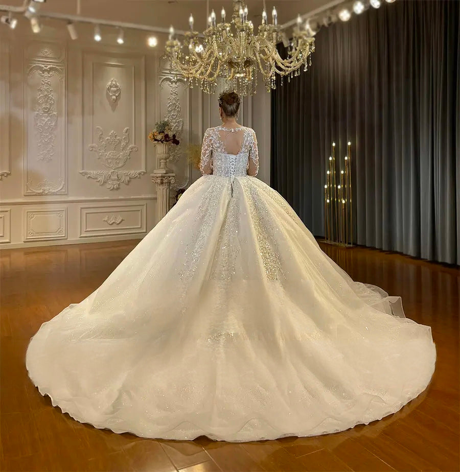 Rear view of a luxury wedding gown highlighting the sophisticated lace-up corset back, sheer illusion lace sleeves, and the beginning of a massive shimmering train.