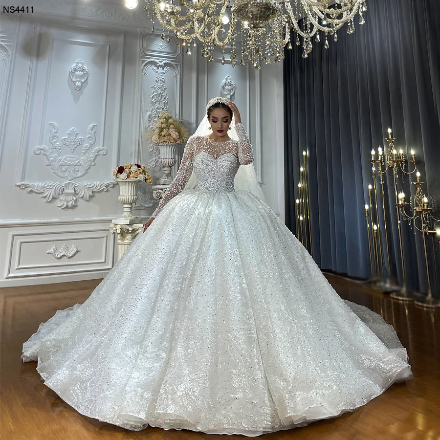 Luxury long sleeve ball gown wedding dress featuring heavy pearl embellishments and an illusion neckline shown on a bride in a grand showroom.