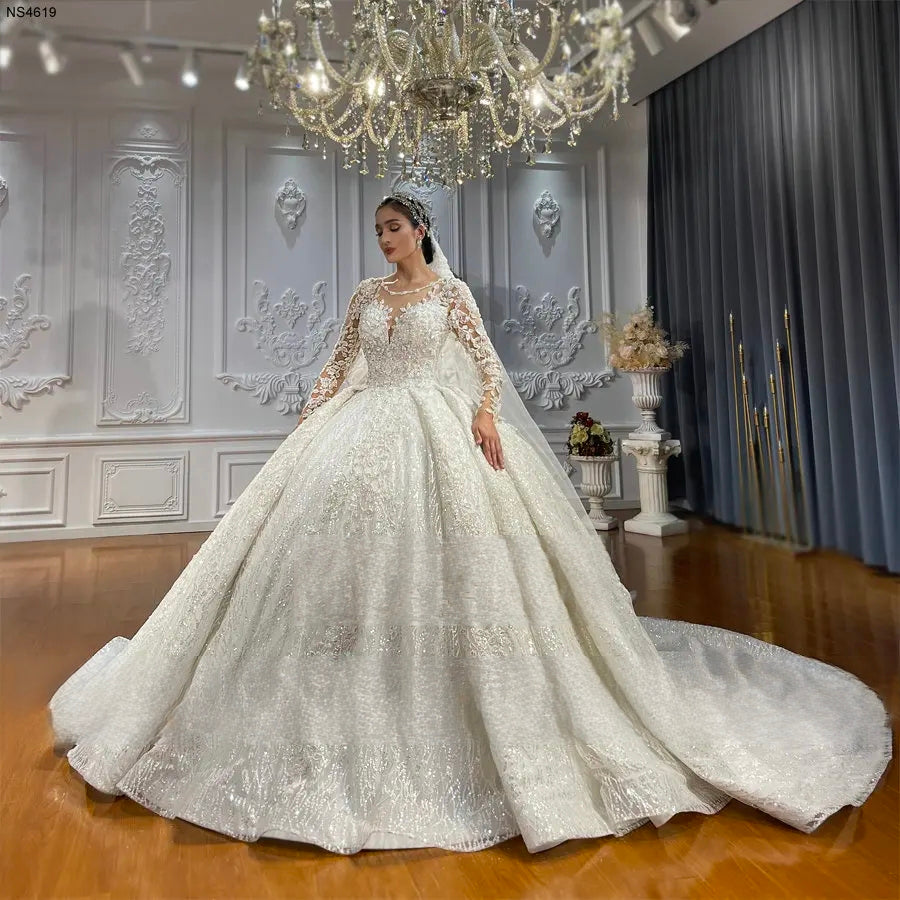 Stunning front view of a sparkling long sleeve ball gown bridal dress, highlighting the regal silhouette and the grand scale of the shimmering tulle skirt under a crystal chandelier.