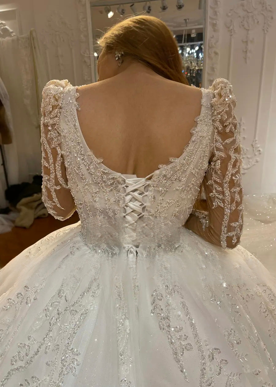Close-up detail of the back of an elegant ivory bridal gown showing an intricate lace-up corset closure and delicate floral embroidery on sheer long sleeves.