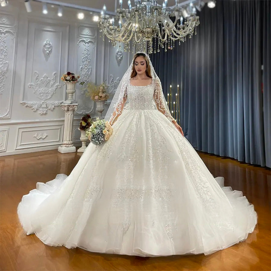 Front-facing view of an exquisite long sleeve lace bridal gown with a full silhouette and intricate crystal embellishments under a chandelier.