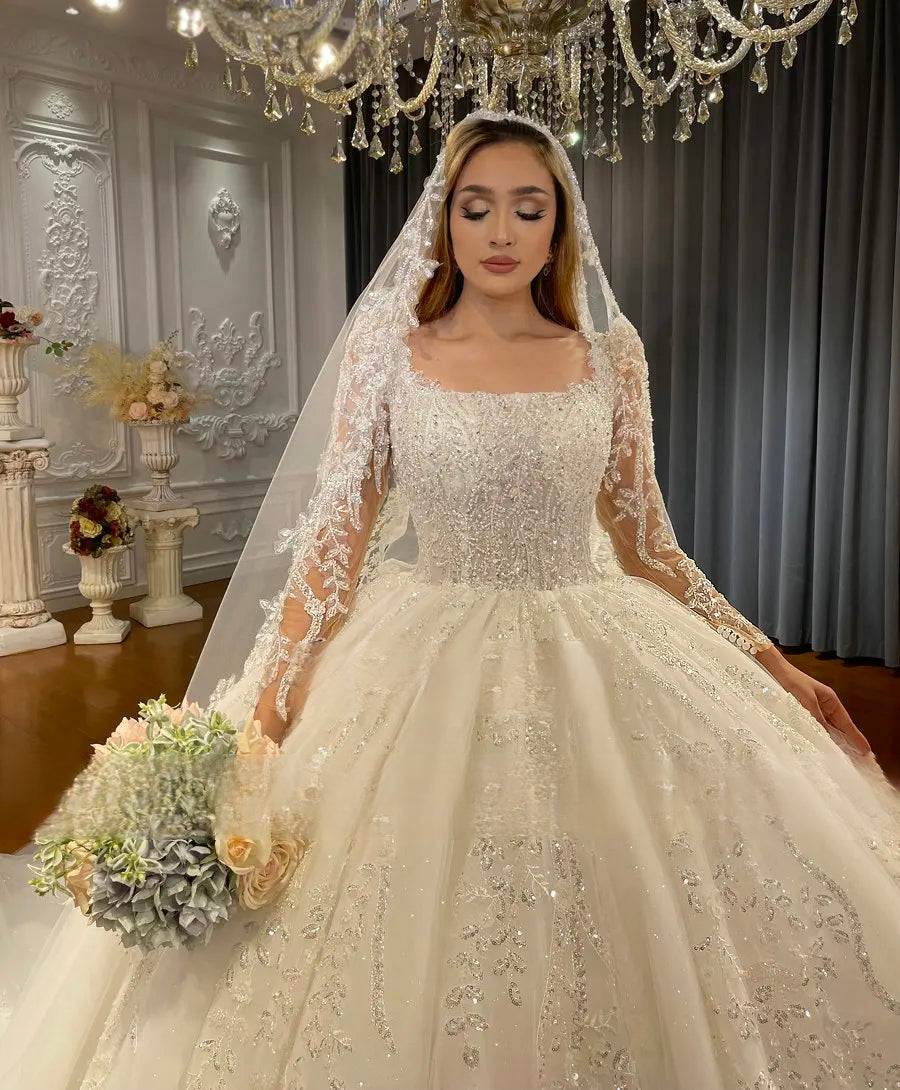 Medium shot of a luxury wedding dress bodice showcasing high-quality lace detailing, a square neckline, and hand-beaded shimmer on long sheer sleeves.