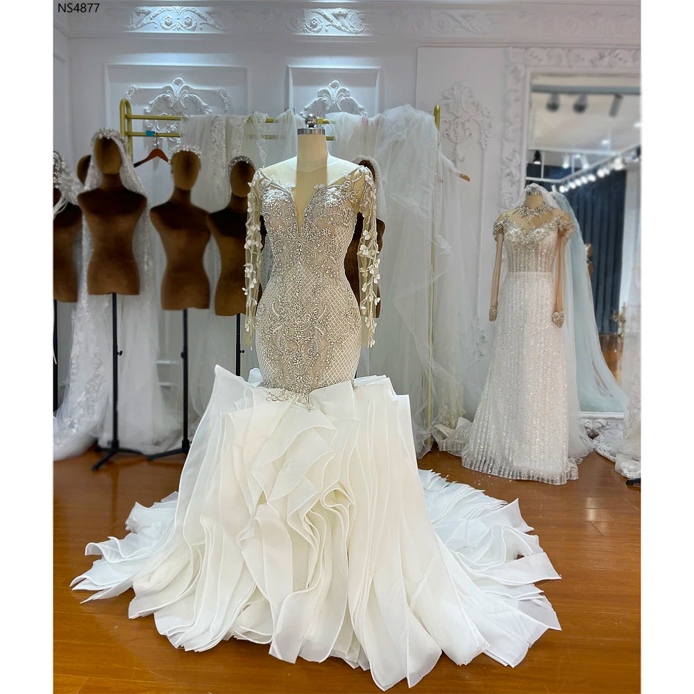 Mannequin display of a beaded mermaid bridal dress with an illusion neckline, long sleeves with 3D floral accents, and a large ruffled skirt.