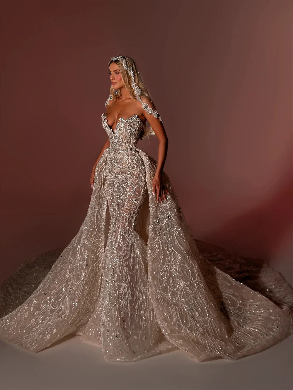 Angled front shot of an opulent crystal-beaded mermaid wedding gown, emphasizing the sculptural sweetheart neckline and layered depth of the detachable lace overskirt.