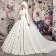 A bride wearing a luxury Muslim A-line wedding dress with long sleeves, high neck, tulle, and intricate lace applique beading, posing in front of a floral backdrop.