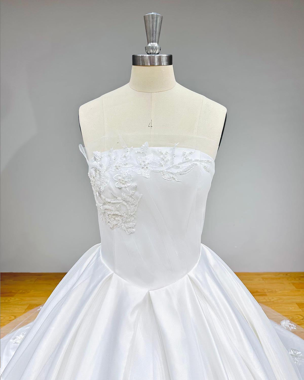 Close-up front bodice detail of a luxury ball gown wedding dress on a mannequin, emphasizing the smooth satin fabric, elegant pleated waistline, and beautiful asymmetrical floral lace appliques.