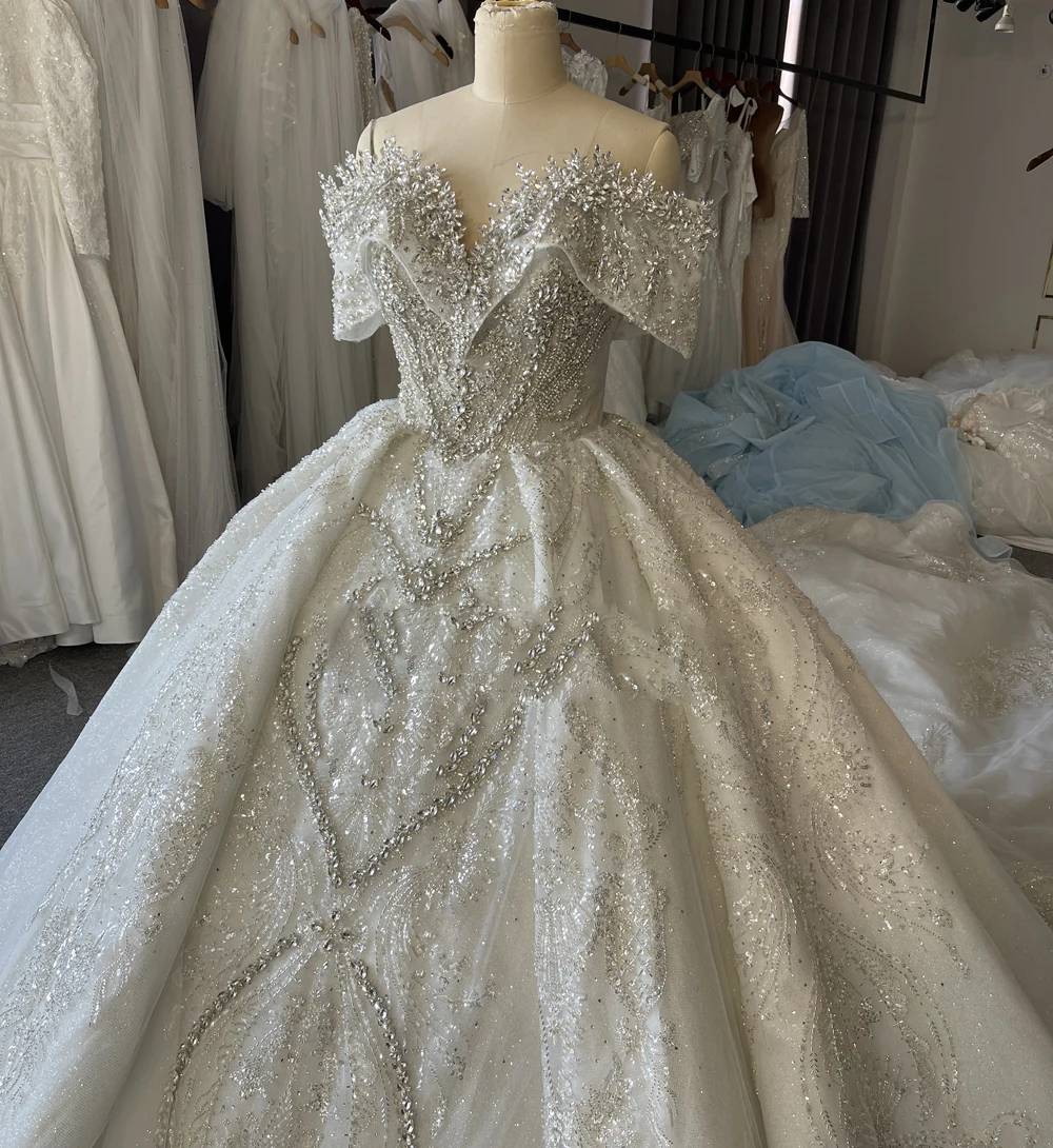 Angled close-up of the luxury off-the-shoulder bridal gown on a mannequin, showcasing the intricate beadwork, delicate sleeves, and voluminous sparkling skirt.