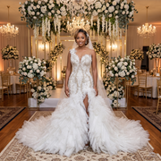A bride stands at an elegant indoor wedding reception, wearing a luxury off-the-shoulder mermaid wedding dress with an intricate beaded bodice, a high front slit, and a voluminous ruffled skirt.