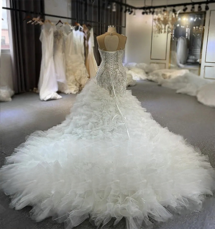 Back view of a beaded mermaid bridal gown showing an elegant heart-shaped keyhole back, corset lace-up closure, and a voluminous tiered ruffle cathedral train.