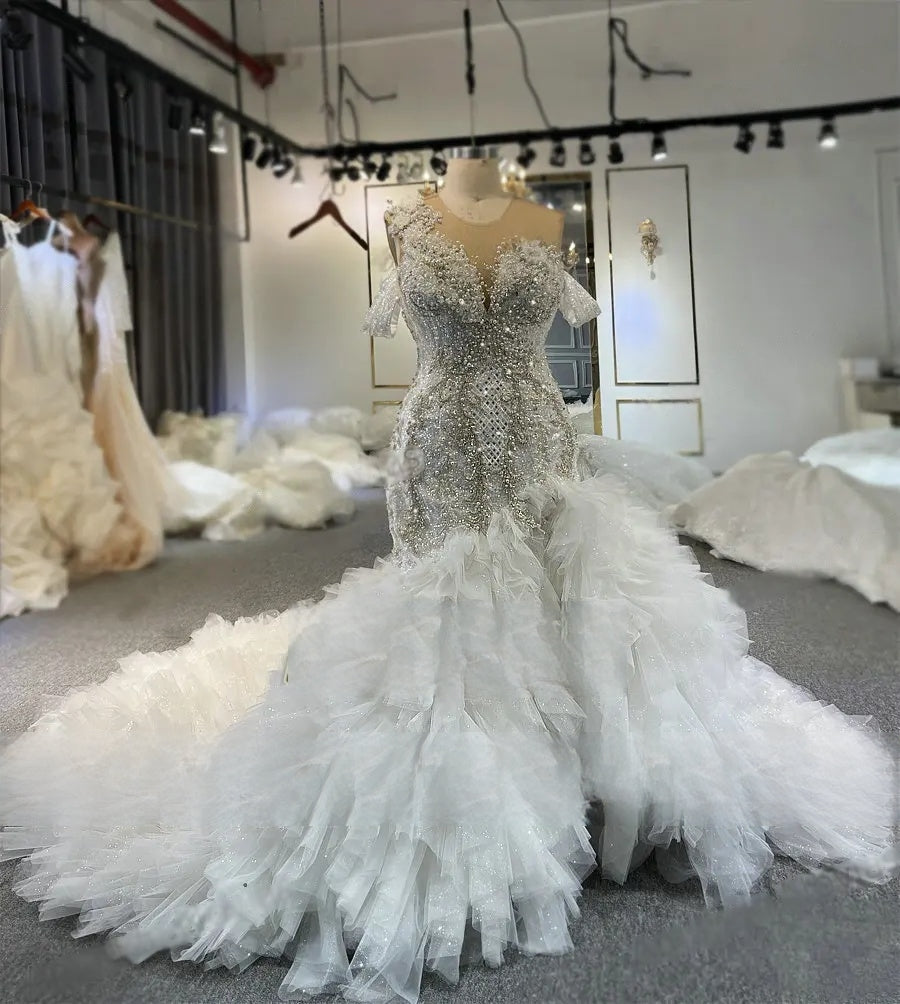 Angled view of an off-the-shoulder luxury wedding dress highlighting the shimmering beaded bodice and a tiered organza ruffle mermaid skirt with a daring front slit.