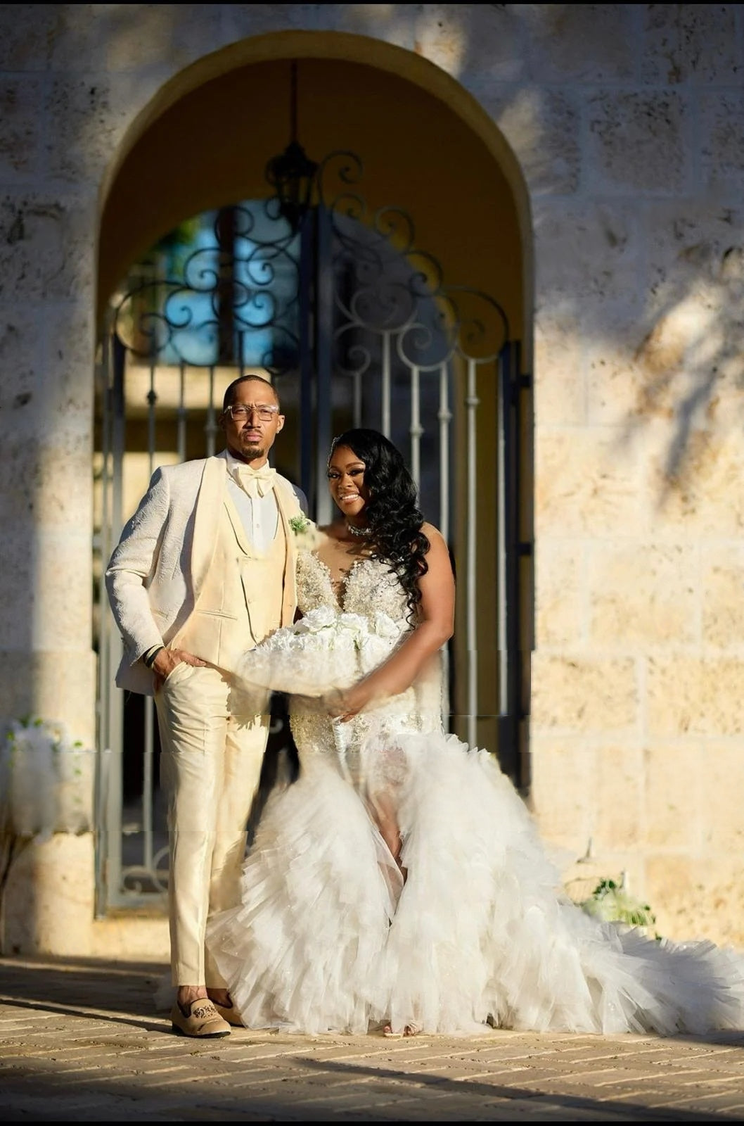 Real bride wearing the luxury mermaid wedding dress with a high slit and ruffled skirt, posed with the groom in a classic outdoor wedding portrait.