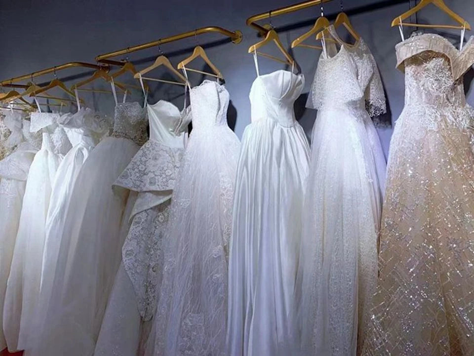 Assortment of premium white and champagne princess wedding gowns with diverse necklines and sequined fabrics displayed on golden hangers in a bridal boutique.