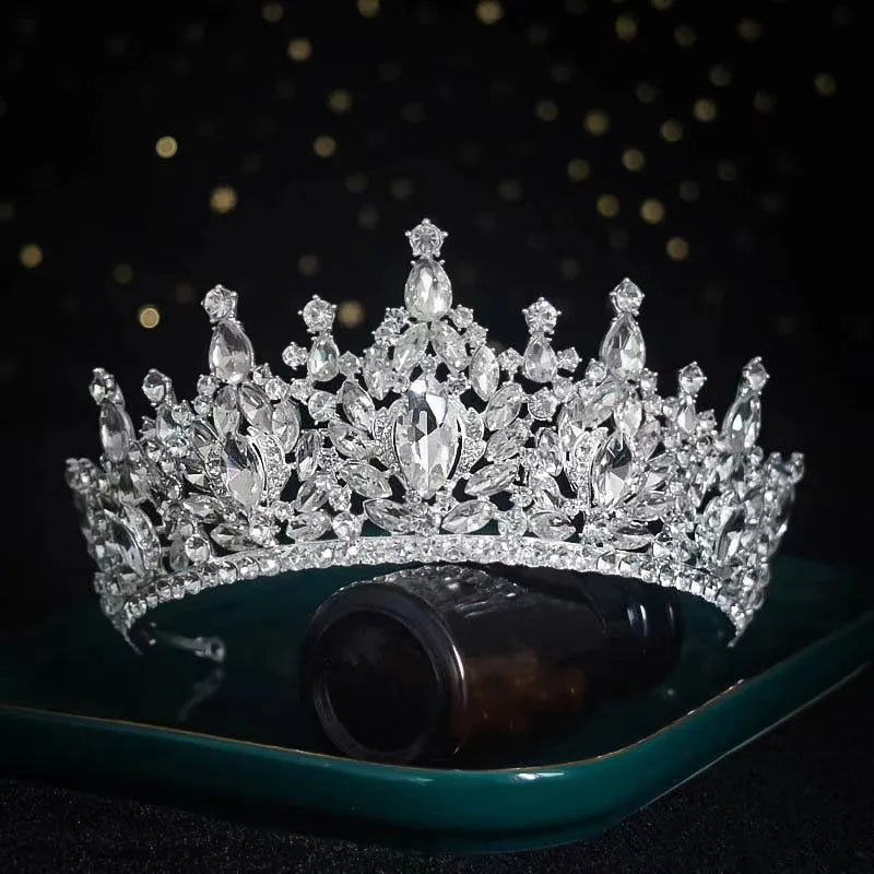 A sparkling luxury bridal tiara crown with marquise-cut rhinestones displayed over a small amber bottle on a dark tray.