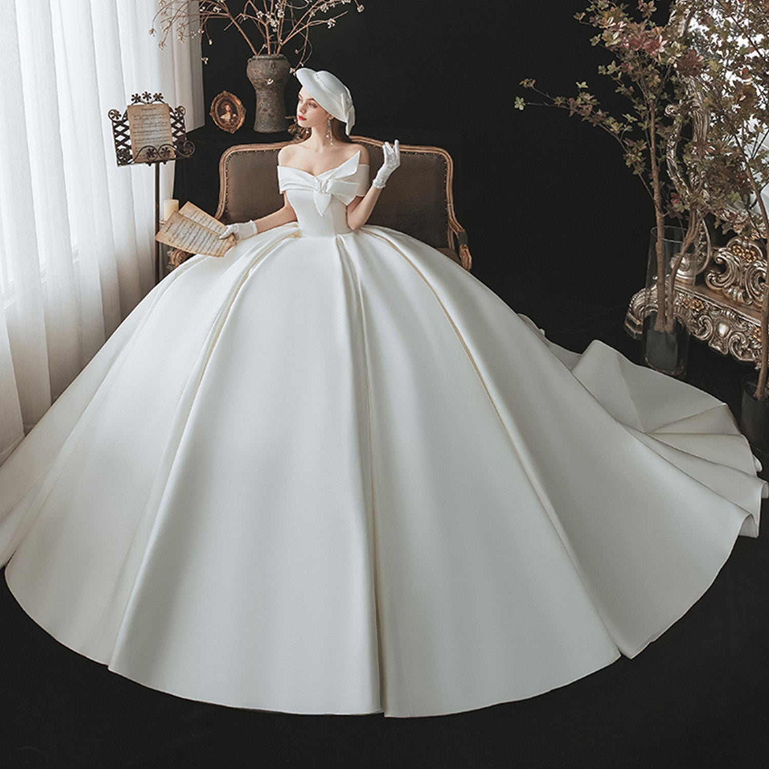 High-fashion bridal portrait of a woman seated in a vintage room wearing an expansive satin ball gown dress with matching bridal gloves and a chic white fascinator hat.