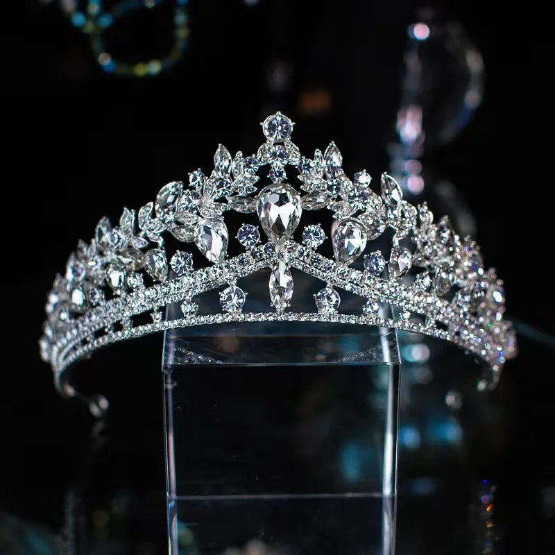 A stunning crystal pageant crown with a central teardrop gemstone and leaf-patterned rhinestone details, displayed on a glass block.