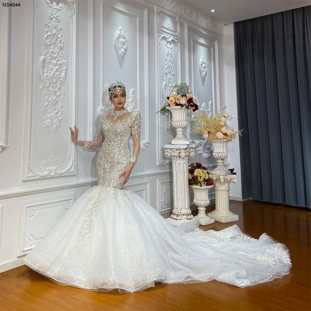 Full-length view of a woman modeling a luxury white mermaid wedding dress featuring heavy crystal beading, a high neckline, and a voluminous shimmering tulle train.