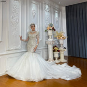 Full-length view of a woman modeling a luxury white mermaid wedding dress featuring heavy crystal beading, a high neckline, and a voluminous shimmering tulle train.
