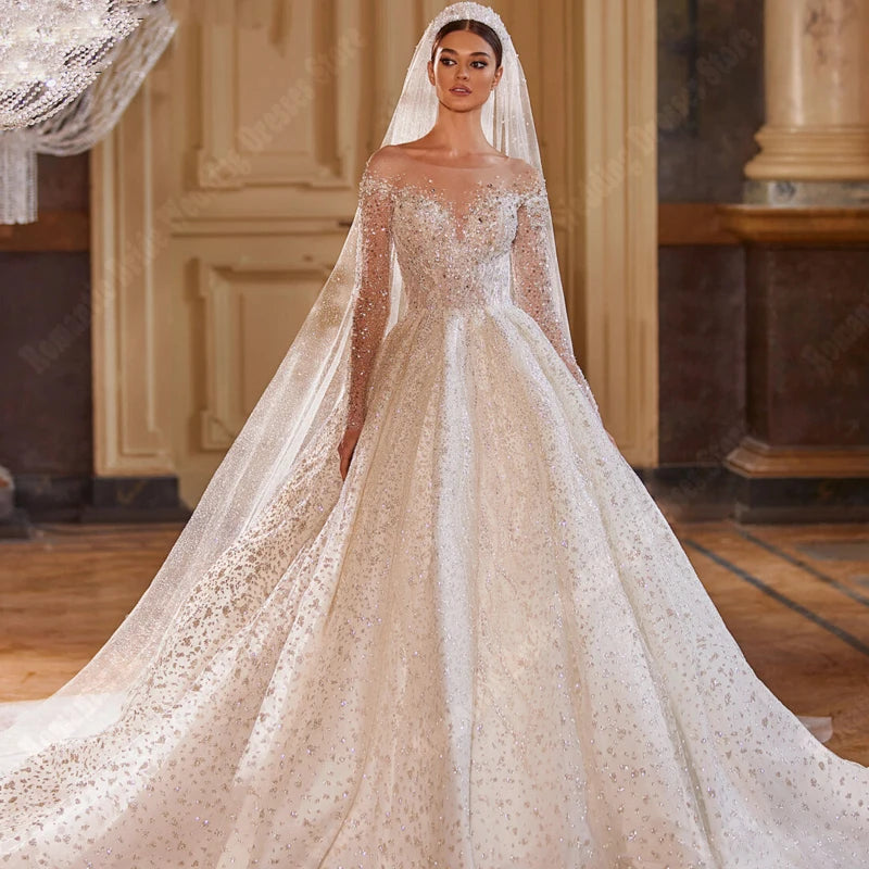 A full-length view of a radiant bride in a V-neck, long-sleeve sequin bridal gown with an A-line skirt and sweeping train, captured in an elegant interior.
