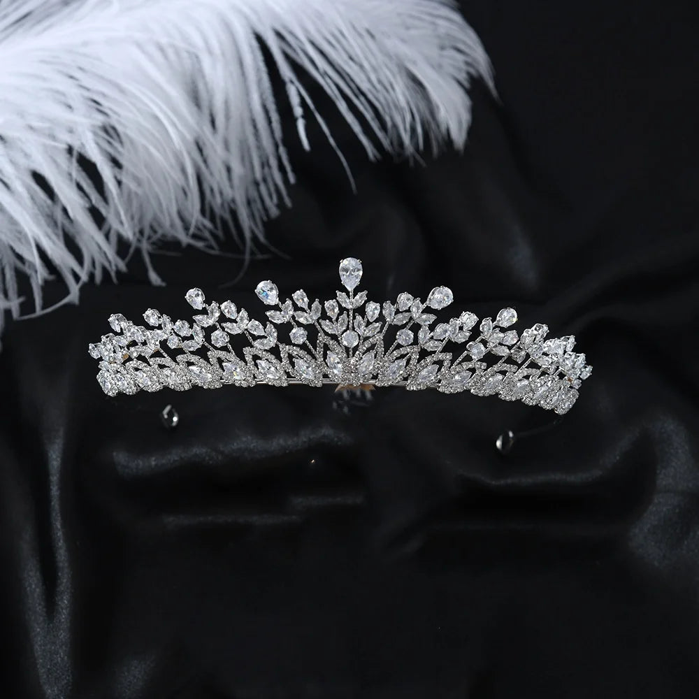 A delicate silver zircon party headpiece crown resting on a black silk satin fabric next to a white feather.