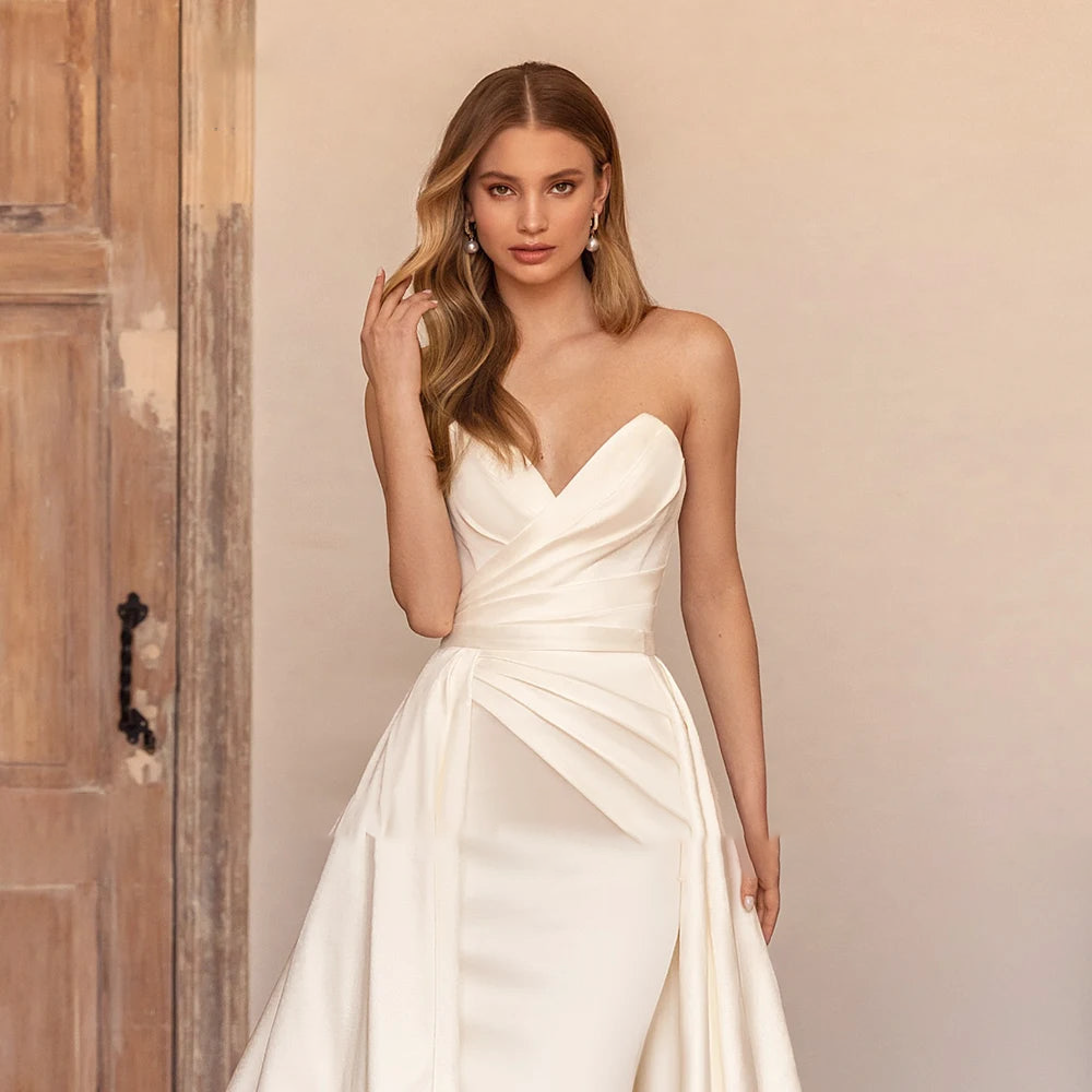 A close-up portrait of a blonde bride posing in a simple, strapless satin mermaid wedding gown with a refined sweetheart neckline.
