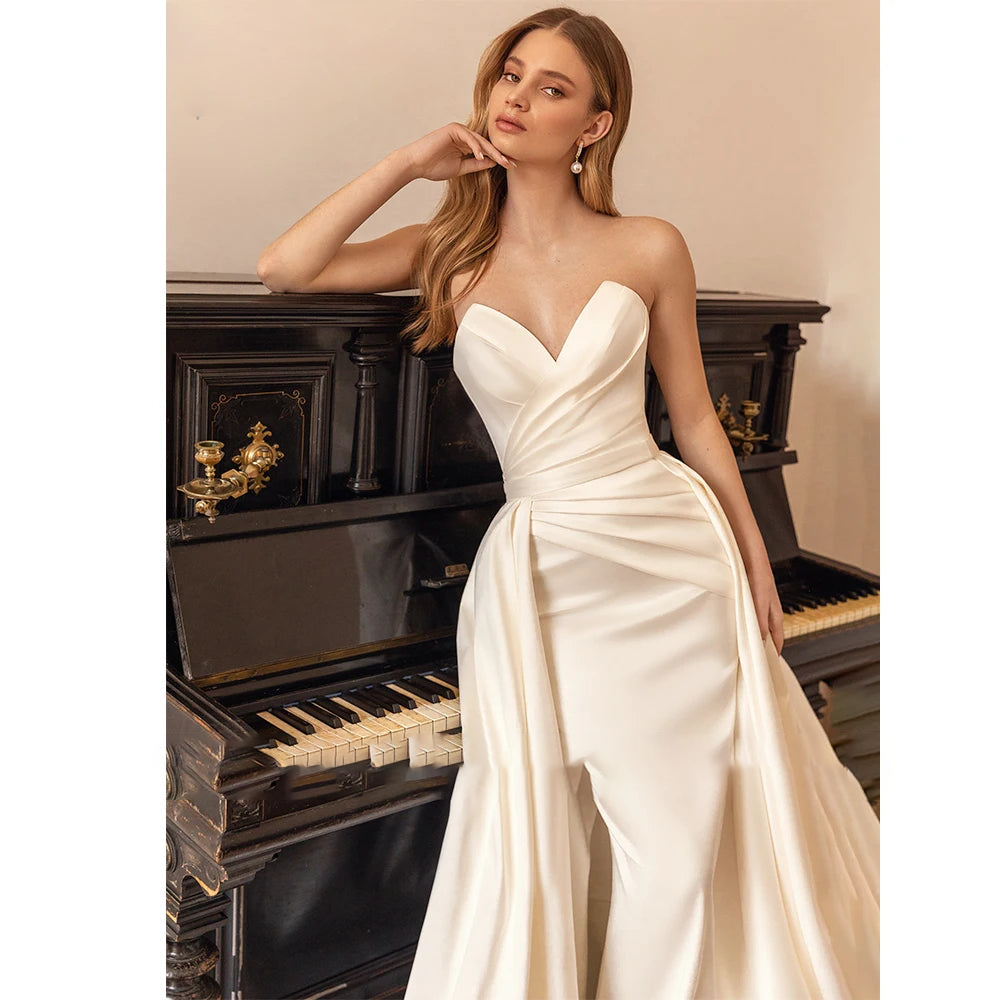 A bride posing beside a classic piano wearing a sweetheart satin mermaid wedding dress with a dramatic detachable train.