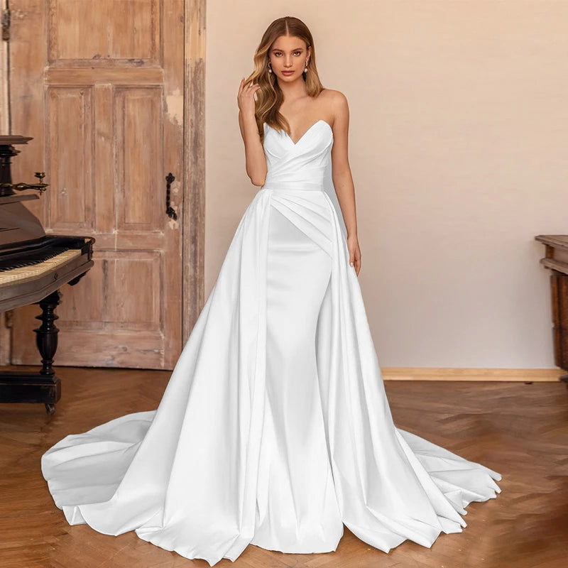 A full-length portrait of a bride modeling a simple and elegant sweetheart satin mermaid bridal gown with a detachable train.