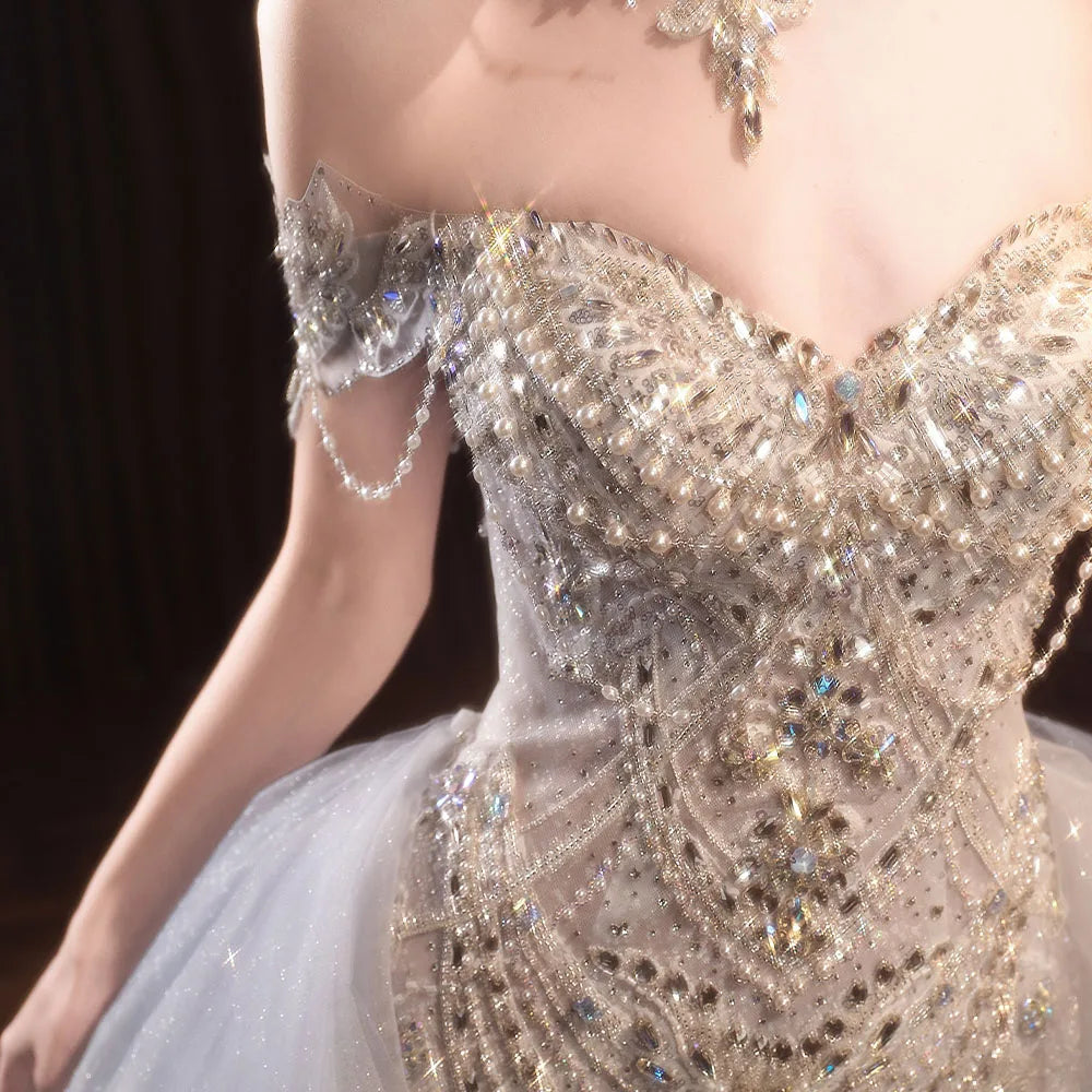 Extreme close-up of a luxury bridal gown bodice, highlighting the dense patterns of iridescent crystals, pearls, a sweetheart neckline, and a draped pearl-chain off-the-shoulder strap.