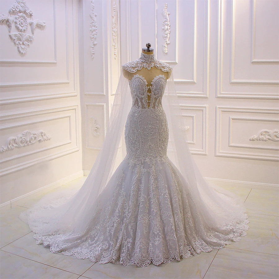 Three-quarter front view of a strapless sweetheart mermaid bridal gown with intricate lace embroidery and a sheer high-neck cape attachment.