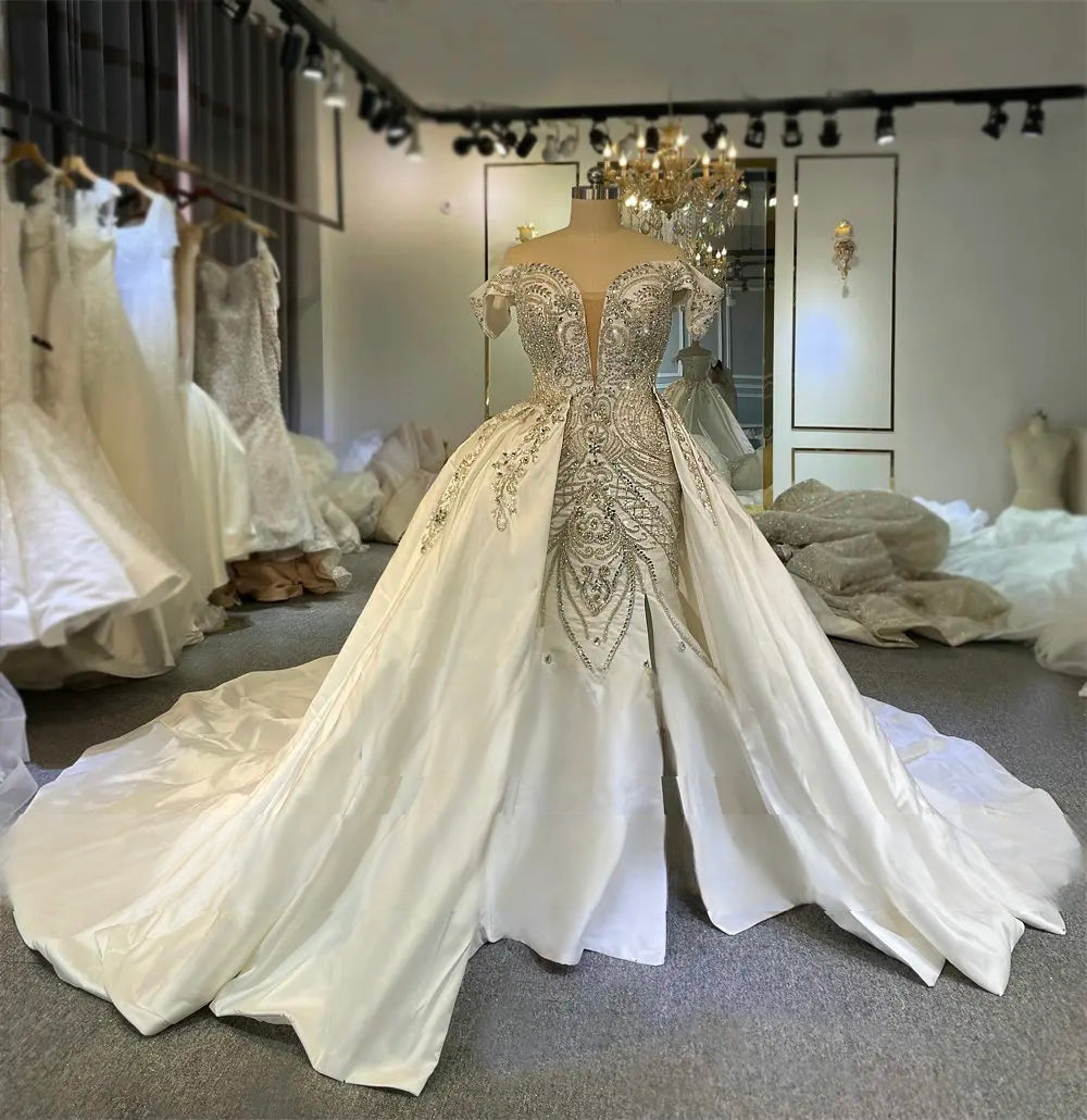 Showroom display of an elegant beaded wedding dress with a 2-in-1 design, featuring a fitted mermaid skirt and a grand detachable Mikado satin train under a chandelier.
