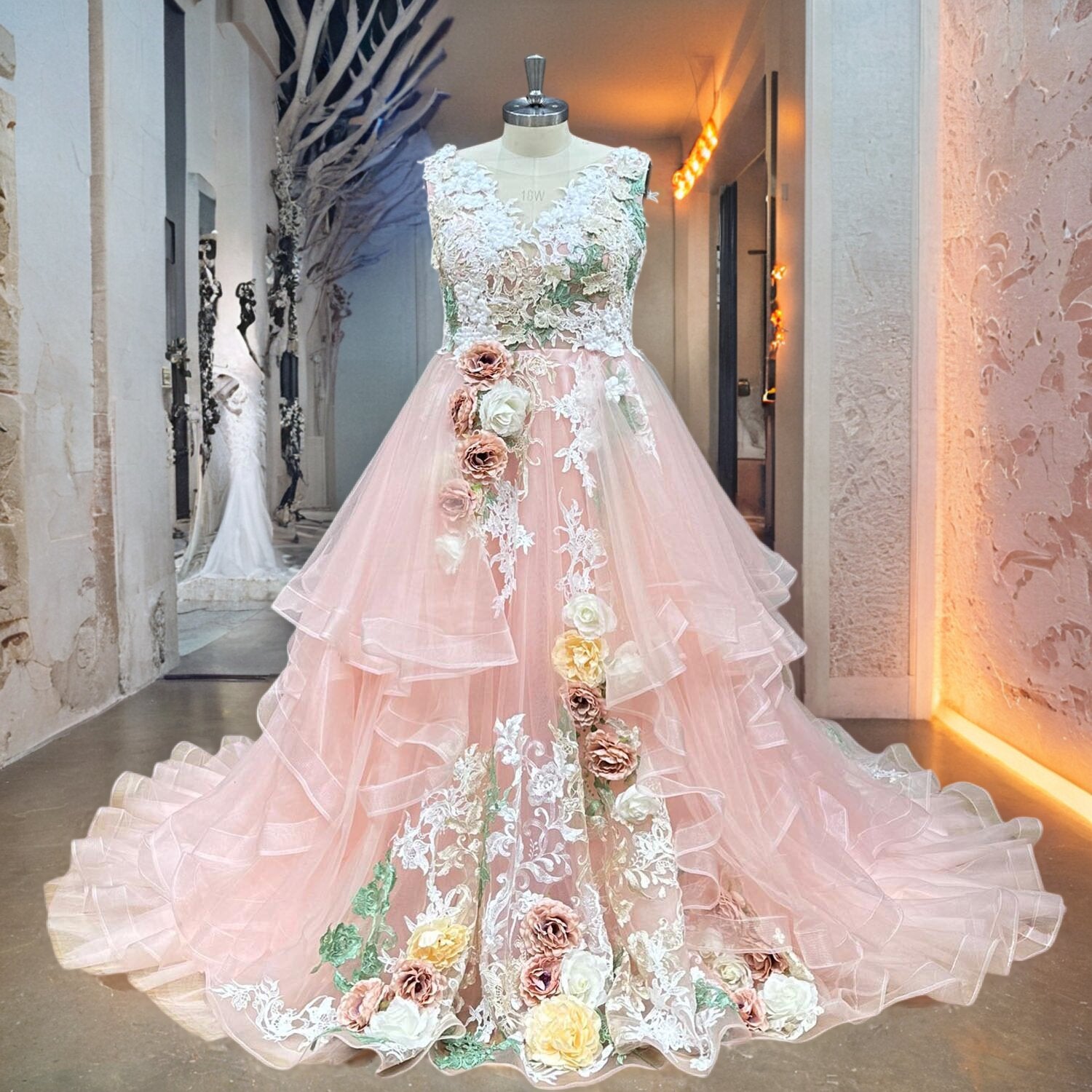 Whimsical pink floral bridal gown with a structured lace bodice and a layered tulle waterfall skirt featuring scattered 3D flower accents, shown on a mannequin in a designer showroom.