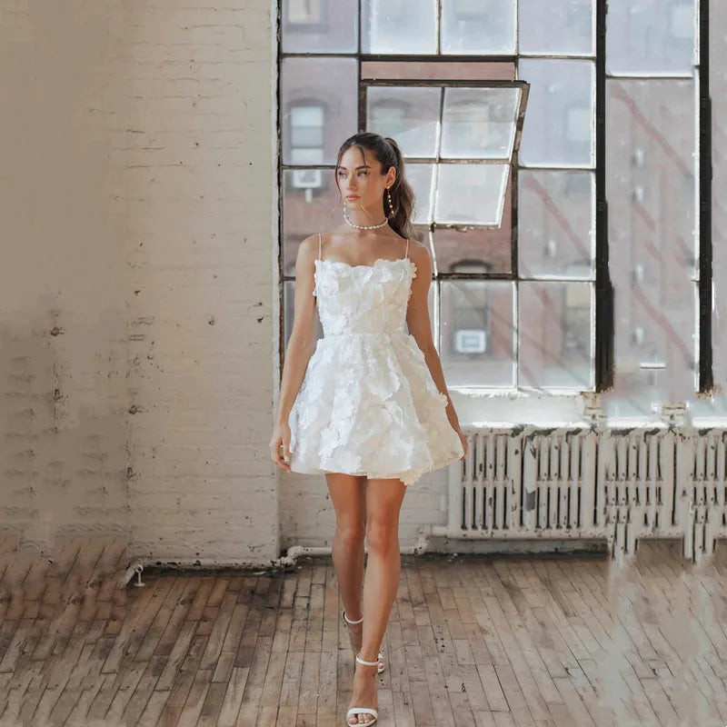 Modern short A-line wedding dress featuring thin spaghetti straps, a fitted bodice covered in 3D textured floral appliques, and a flirty volumetric mini skirt.