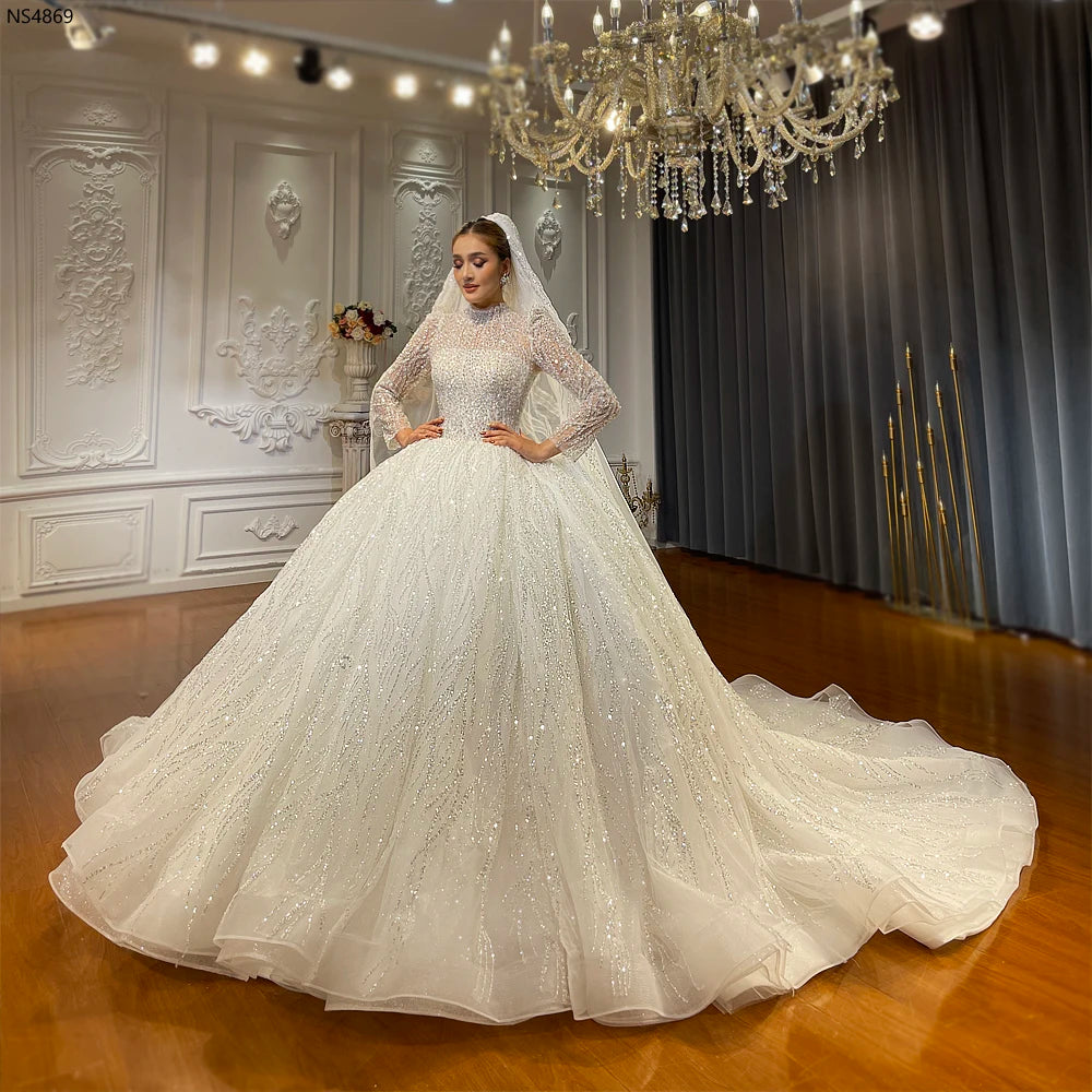 Full length, wide angle shot of a model showcasing a highly embellished, long-sleeved modest Muslim bridal ball gown with a magnificent train, set against an elegant showroom backdrop.