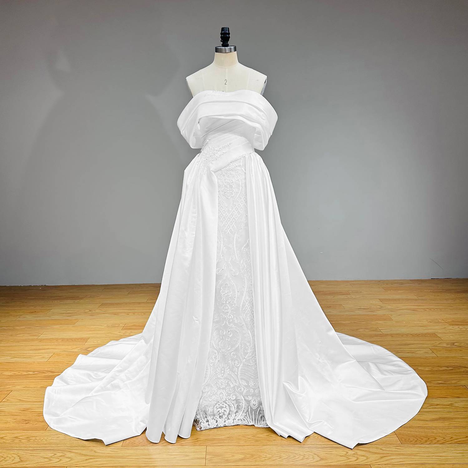 Three-quarter studio view of an architectural white satin wedding dress featuring a modern boat neck, draped portrait collar, and an embroidered lace column skirt.